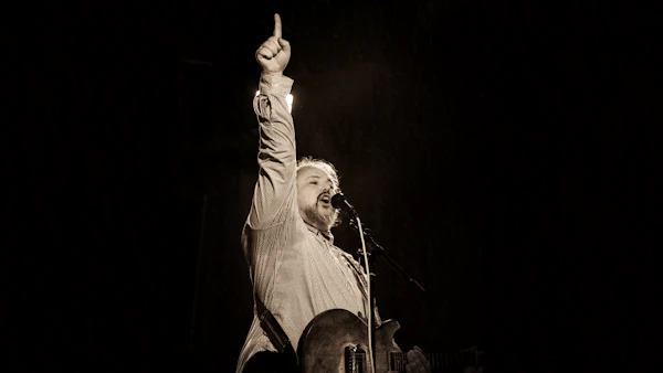 Live on stage, singing into a microphone with a guitar and one arm raised.